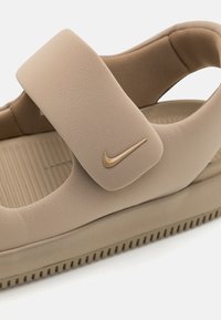 Beżowe obuwie z teksturowaną cholewką, regulowanym paskiem i złotym logo Nike. Posiada żłobioną podeszwę, zapewniającą lepszą przyczepność i komfort.