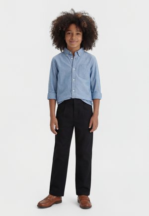 Barn med krøllete hår iført en lyseblå skjorte med knapper, svarte bukser og brune sko, står foran en hvit bakgrunn.