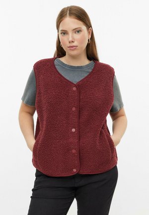 Femme portant un gilet polaire bordeaux boutonné sur un t-shirt gris à manches courtes, les mains dans les poches du gilet, debout devant un fond blanc.