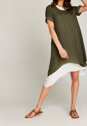 Femme portant une tunique verte à manches courtes superposée sur une robe blanche asymétrique, associée à des sandales plates marron, posant devant un fond uni.
