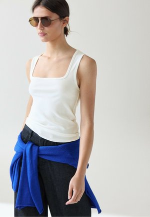 Frau trägt braune Sonnenbrille, weißes ärmelloses Oberteil, schwarze Hose und einen hellblauen Pullover um die Taille gebunden.