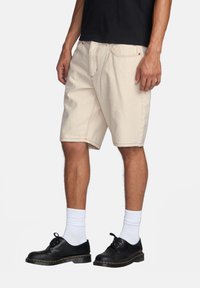 Shorts color crema di lunghezza al ginocchio con cuciture a contrasto, abbinati a scarpe nere e calze bianche. Design semplice, materiale di cotone.