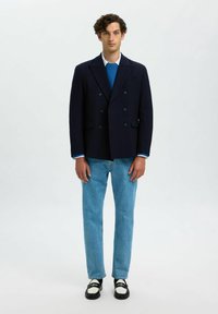 Jeune homme aux cheveux bouclés portant un blazer noir croisé, un pull bleu, un jean bleu clair et des mocassins noir et blanc, se tenant sur fond blanc.