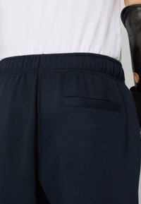 Mørkeblå joggebukser med elastisk midje, med en liten baklomme og jevn tekstur. Inkluderer kontrastfargede stikninger.