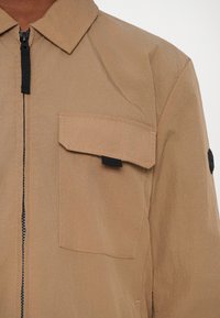 Beige Außenjacke mit spitzem Kragen, Reißverschluss und einer Brusttasche. Verfügt über einen strukturierten Stoff und schwarze Hardware-Akzente.