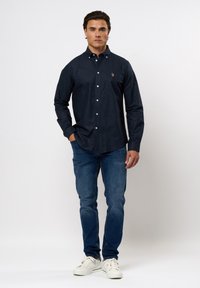 Navy-Blaues Hemd mit Knopfleiste aus Baumwolle, mit langen Ärmeln und kleinem Logo auf der Brust, kombiniert mit blauen Jeans und weißen Sneakers.
