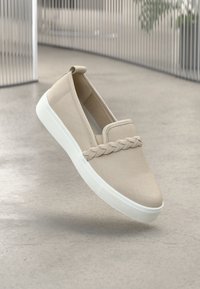 Beige slip-on shoe made of canvas with a braided detail across the front and a white rubber sole. Minimalistic design with elastic sides.