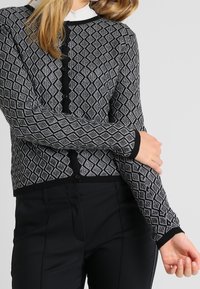 Svartvit mönstrad cardigan med diamantdesign, rund halsringning och knappstängning framtill; åtsittande ärmar med kontrasterande svarta kanter.