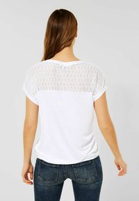 Bílé tričko s texturovaným krajkovým zadním dílem, krátkými rukávy a volnějším střihem, v kombinaci s tmavě modrými džínami.