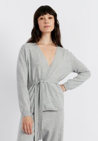 Grijze gebreide omslagtrui met lange mouwen, diepe V-hals en strikceintuurdetail. Zachte textuur, comfortabele pasvorm en geribde manchetten voor extra comfort.