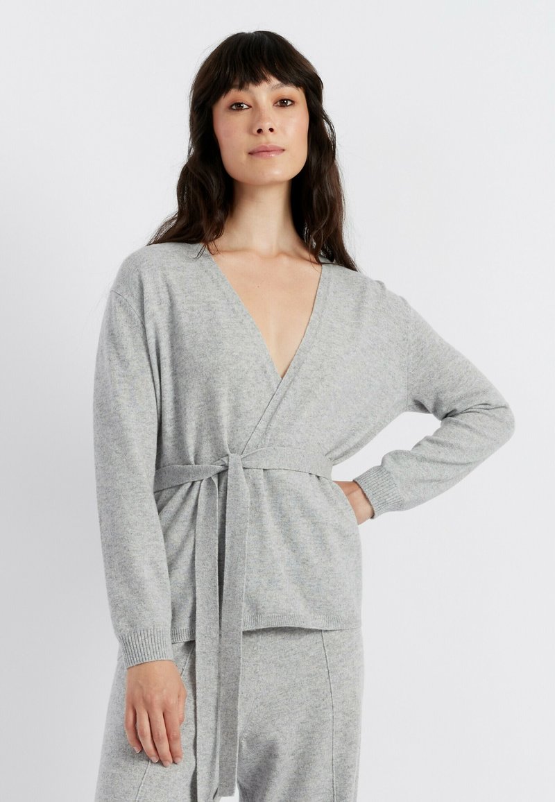 Grijze gebreide omslagtrui met lange mouwen, diepe V-hals en strikceintuurdetail. Zachte textuur, comfortabele pasvorm en geribde manchetten voor extra comfort.