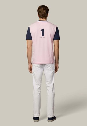 Hombre de pie de espaldas vistiendo una camisa de manga corta rosa y azul marino con el número 1 y el texto "Hackett", pantalones blancos y zapatos oscuros.