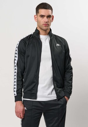 Man draagt een zwarte trainingsjas met rits en bijpassende broek met witte logostrepen over een wit T-shirt, staand tegen een effen achtergrond.