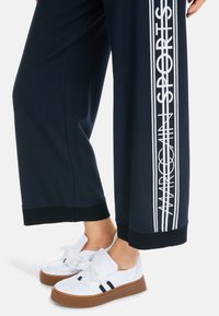 Marineblaue Sweatpants mit weißem Textdesign an der Seite, ausgestattet mit gerippten Bündchen. Kombiniert mit weißen Sneakers mit einer hellbraunen Plateausohle.
