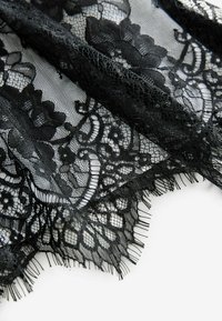 Czarna koronkowa tkanina z kwiatowymi wzorami, z subtelnymi detalami, przejrzystą teksturą i frędzlami, ułożona na jasnym tle.