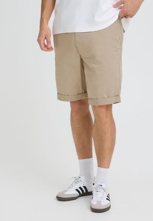 Mand iført beige knælange shorts, hvid t-shirt, hvide sokker og hvide sneakers med sorte striber, stående mod en ensfarvet baggrund.