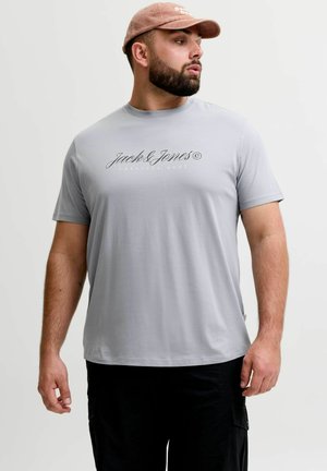 Muškarac u svijetlosivoj majici "Jack & Jones", crnim hlačama i roza bejzbol kapom, gleda lijevo na bijeloj pozadini.