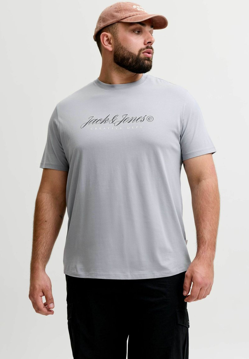 Άνδρας που φοράει ανοιχτό γκρι μπλουζάκι "Jack & Jones", μαύρο παντελόνι και ροζ καπέλο μπέιζμπολ, κοιτάζει προς τα αριστερά με φόντο λευκό.