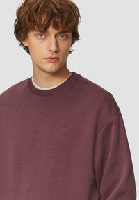 Burgunderroter Pullover mit langen Ärmeln und Rundhalsausschnitt. Verfügt über ein dezentes, auf der Brust gesticktes QS-Logo. Weicher, strukturierter Stoff.