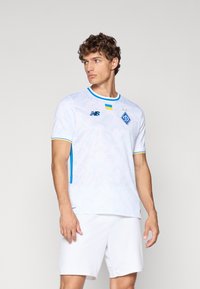 Hvit fotballtrøye med kort erme som har et teksturert mønster, blå detaljer og rund hals. Inkluderer laglogo og gul kant.