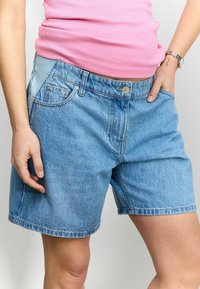 Shorts de mezclilla azul claro con un corte suelto, bolsillos frontales y cierre de botón. La cinturilla presenta una sección de un azul más claro que contrasta.