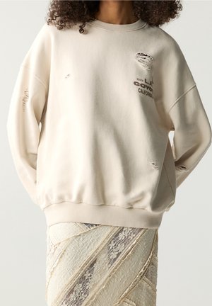 Personne porte un sweat-shirt crème usé avec un texte et une jupe en dentelle crème texturée, les mains cachées dans le dos.