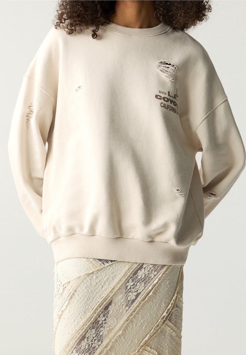 Personne porte un sweat-shirt crème usé avec un texte et une jupe en dentelle crème texturée, les mains cachées dans le dos.