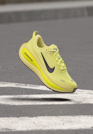 Chaussure de course Nike ZoomX jaune en l'air au-dessus d'un bitume gris avec des marquages routiers blancs et de petites particules de gravier.