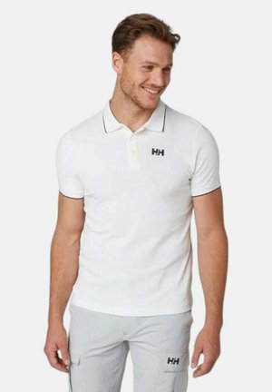 Camisa polo de algodón blanca con cuello, con ribetes negros y un logo bordado en el lado izquierdo del pecho. Combinada con pantalones cargo gris claro.