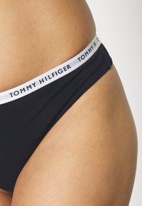 Bas de bikini noir avec une mince taille blanche imprimée de "TOMMY HILFIGER." Texture lisse, coupe classique, montrant une vue de côté.