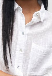 Chemise en lin blanc avec un motif à carreaux texturé, dotée d'une patte de boutonnage devant, d'un col standard et d'une poche poitrine unique. Les boutons sont bleu clair.