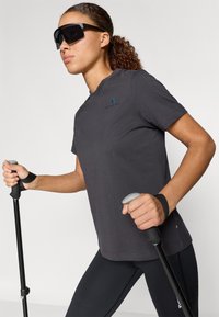 Sötétszürke sportpóló rövid ujjakkal, egy kis logóval. Fekete leggingsekkel és ergonomikus markolatú túrabotokkal párosítva.