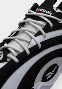 Κοντινό πλάνο μαύρου και λευκού αθλητικού παπουτσιού Reebok με υφασμάτινο πλέγμα, χοντρές λευκές κορδόνια και λογότυπα της μάρκας στη γλώσσα και την πλαϊνή πλευρά.