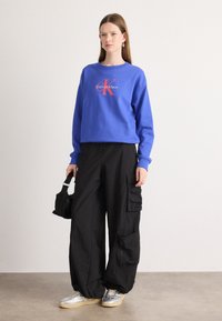 Felpa blu royal con collo rotondo e logo "Calvin Klein"; pantaloni cargo neri con tasche; sneakers argento con suole beige; borsa nera.