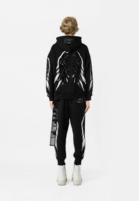 Sudadera negra con diseños de alas en la espalda, combinada con pantalones de chándal negros con etiquetas. Ambos elementos hechos de tela suave, con costuras detalladas.