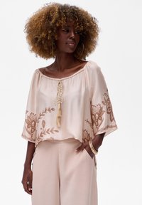 Beige geborduurde blouse met wijde mouwen, decoratieve franjes, bloemenpatronen in donkerdere tinten, gecombineerd met bijpassende broek.