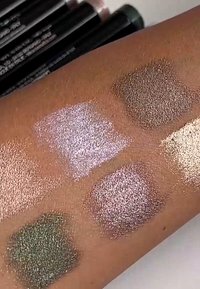 Próbki pięciu metalicznych odcieni cieni do powiek na skórze: zielony, różowy, srebrny, brązowy i złoty, prezentujące różnorodne faktury i wykończenia.
