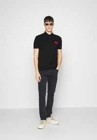 Polo noir à manches courtes, arborant une petite broche logo rouge. Porté avec un jean sombre et des baskets blanches. Design minimaliste.