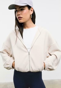 Lichtbeige rits-hoodie met een ontspannen pasvorm, met opgerolde mouwen en een capuchon, gedragen met een wit T-shirt en marineblauwe legging.