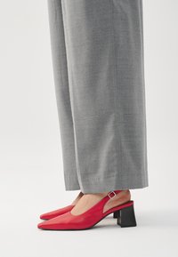 Red leather slingback shoes with a pointed toe, a block heel, and a side buckle detail, paired with gray tailored pants.