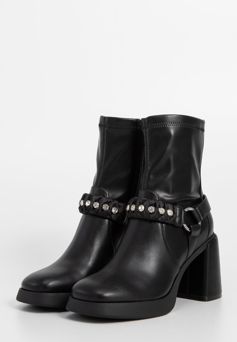 Botas de tobillo de cuero negro con un alto tacón cuadrado, que cuentan con una correa trenzada adornada con tachuelas plateadas. Superficie lisa con un cierre de cremallera.