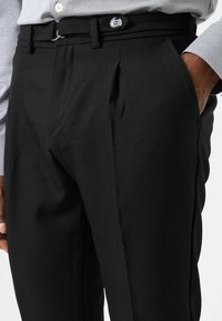 Zwarte maatwerkbroek met zijzakken, geplooide voorkant en een platte tailleband. Gladde stoftextuur met een gestroomlijnde silhouet.