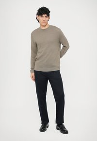 Grauer strukturierter Pullover mit rundem Ausschnitt, kombiniert mit dunkler Hose und schwarzen Schuhen. Der Pullover hat ein dezentes Muster und gerippte Bündchen.