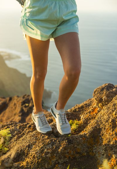 Chaussures de sport gris clair avec des accents blancs, dotées d'un dessus en maille et d'un système de laçage, portées avec un short vert menthe sur un terrain rocailleux.