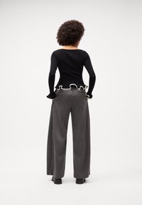 Schwarzes Langarmoberteil mit weißen Rüschendetails, kombiniert mit hoch taillierten grauen Streifen-Hosen mit weitem Bein und klobigen schwarzen Schuhen.