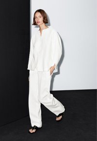 Vrouw in een losse witte blouse en wijde witte broek wandelt over een zwarte vloer tegen een zwart-wit muur.