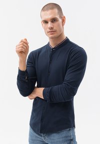 Tummansininen pitkähihaista nappipaitaa, jossa on pystykaulus, punainen ja sininen yksityiskohta raita sekä kapeat hihansuut nappikiinnityksellä.