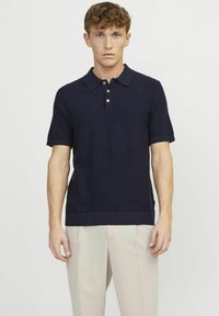 Polo shirt lavorato a maglia di colore navy con maniche corte, alla moda con una chiusura a tre bottoni, colletto e orlo a costine. Indossato con pantaloni di colore chiaro.