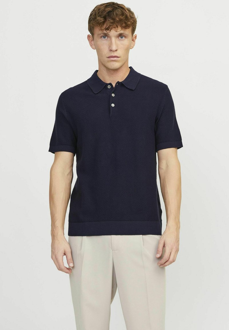 Polo shirt lavorato a maglia di colore navy con maniche corte, alla moda con una chiusura a tre bottoni, colletto e orlo a costine. Indossato con pantaloni di colore chiaro.