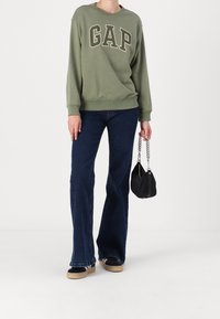Olivově zelená mikina s logem "GAP", spárovaná s tmavomodrými rozšířenými džíny. Drží malou černou kabelku s řetízkovým popruhem. Neformální outfit.